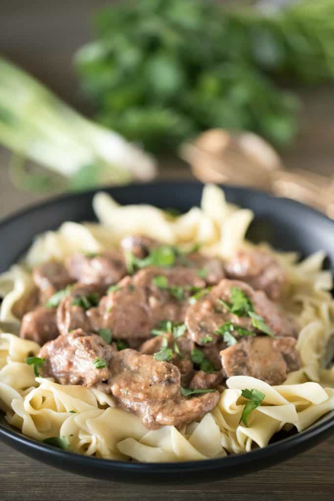 Black plate with cooked egg noodles and topped with beef stroganoff. Green onions and parsley in the background.