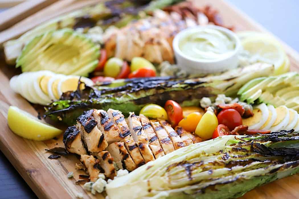 Side view of deconstructed Cobb Salad on platter with marinated and grilled chicken, grilled romaine, cherry tomatoes, avocado, blue cheese and bacon.
