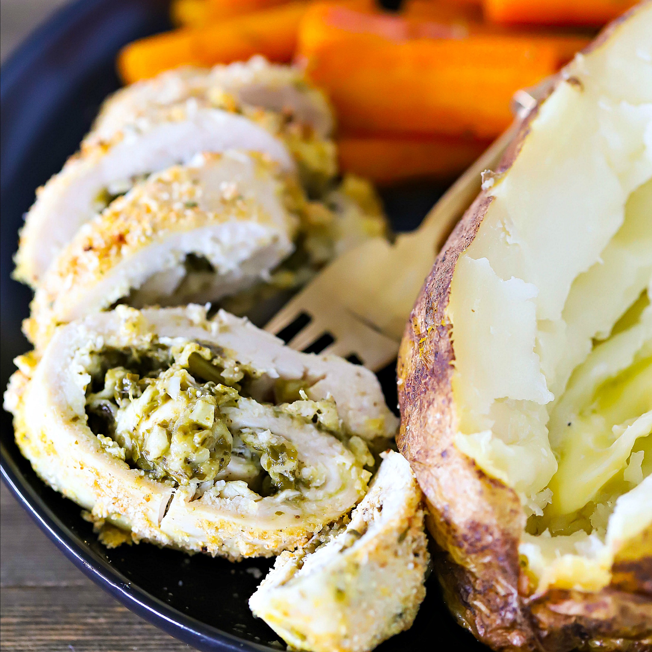 Sliced Pesto Stuffed Chicken on a black plate with a side of carrots and baked potato and a fork.
