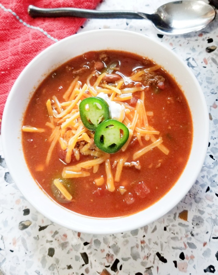 Overhead photo of a white bowl filled with chili, topped with jalapeño and cheese.